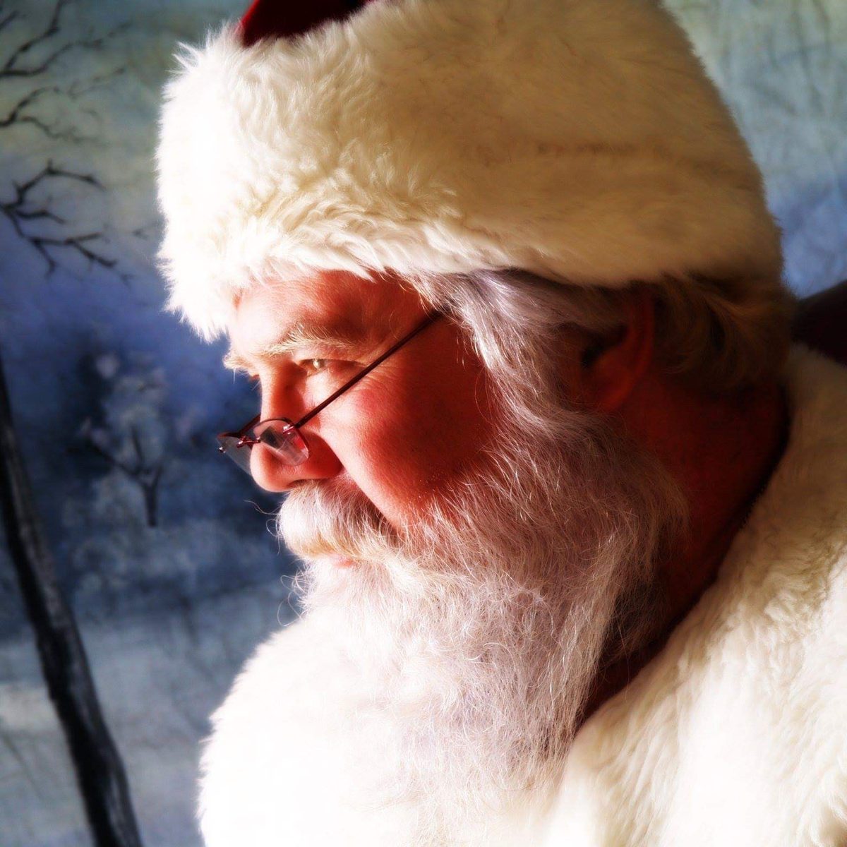 Santa John in profile, in his red suit and hat, against a painted winter backdrop