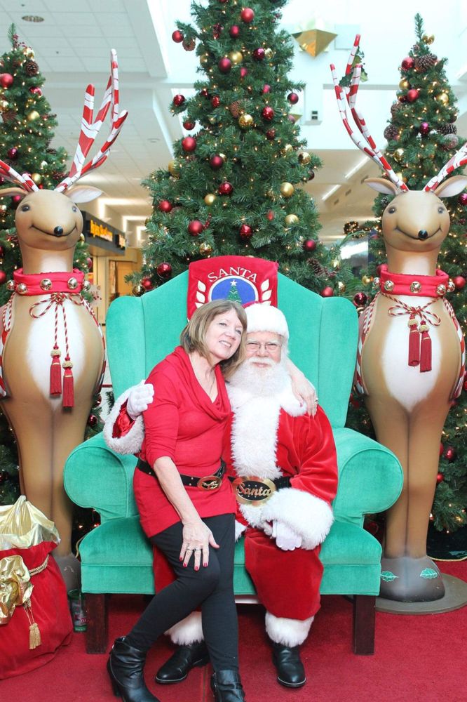 Santa John on a green chair between two reindeer figures, sharing a photo with a visitor